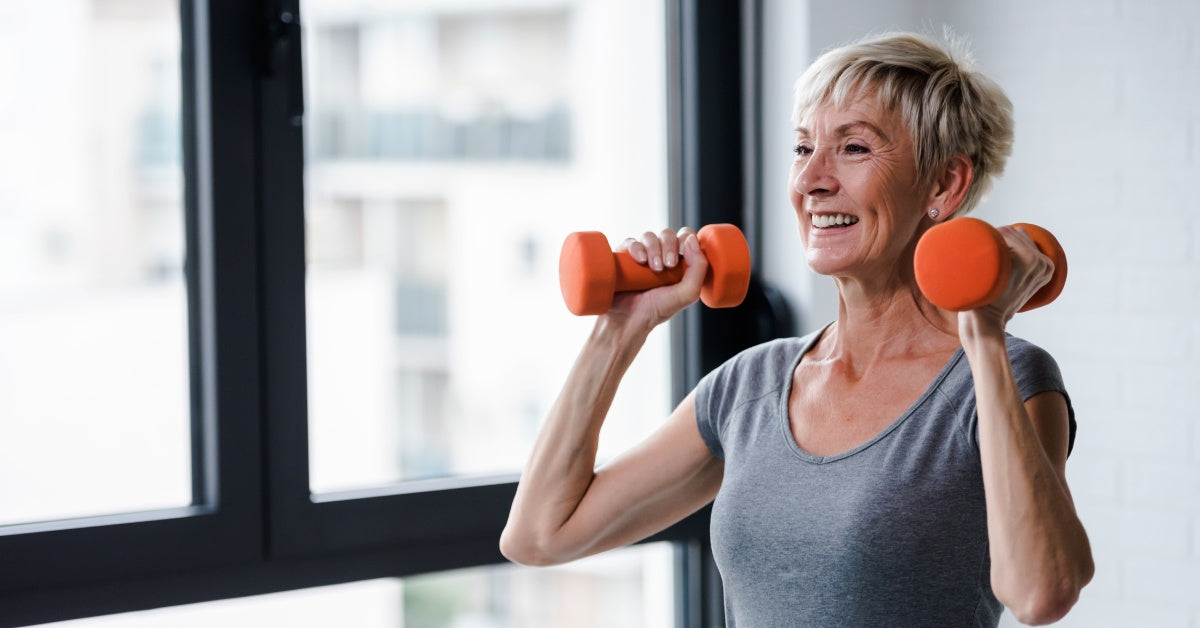 An older woman wearing a gray shirt is smiling and holding two orange dumbbells near her shoulders.